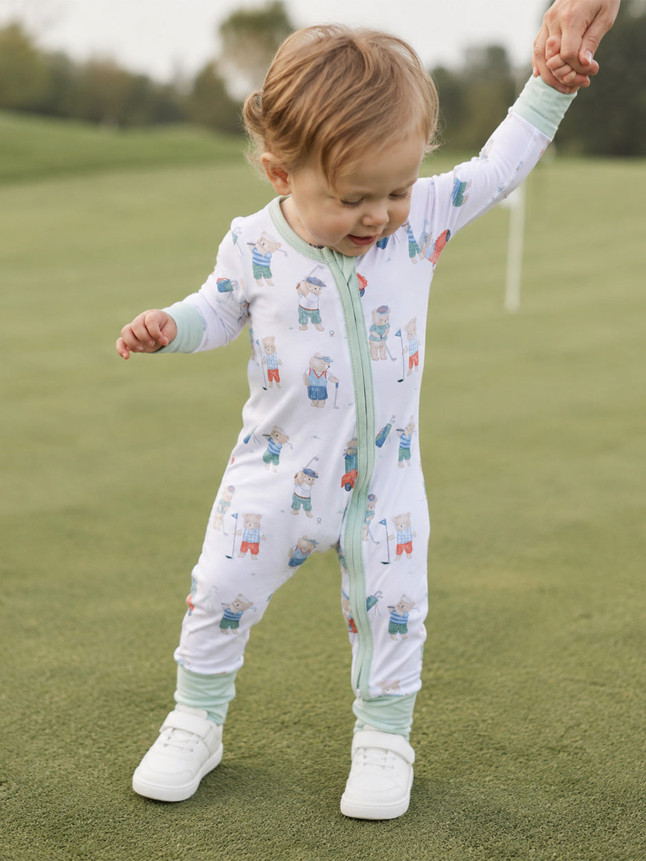 Toddler in playful golf-themed onesie, joyfully walking on a green, held by an adult's hand.