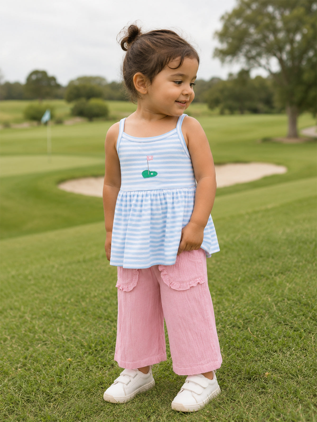 Smiling child in a striped blue top and pink pants stands on grass, with a golf course in the background.