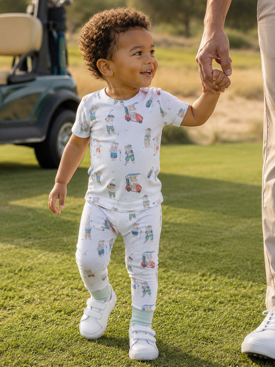 Smiling toddler in golf-themed pajamas walking hand-in-hand with an adult on a grassy field.