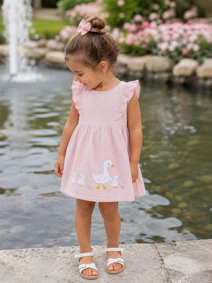 Young girl in a pink dress with duck prints, standing by a pond with flowers and a fountain in the background.