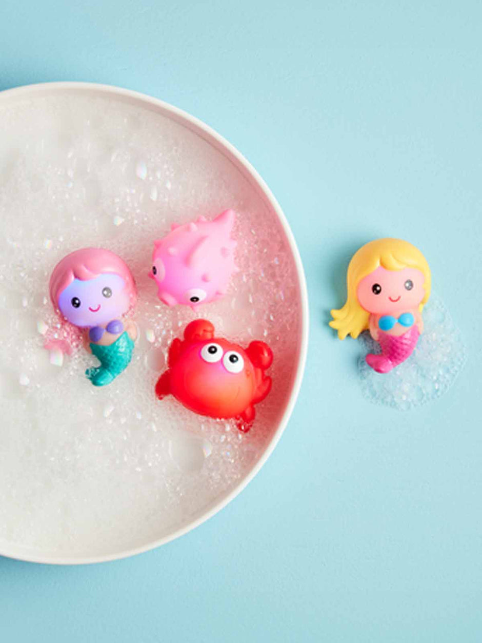 Colorful bath toys, including mermaids and a crab, floating in bubbly water on a light blue background.