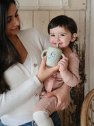 Mother holding smiling baby with a cup in a cozy setting, both enjoying a warm moment together.