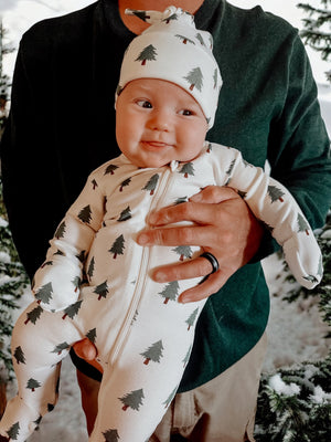 Infant wearing tree-patterned pajamas, smiling while being held by an adult in a snowy forest setting.