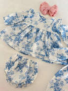 Baby dress and bloomers in white with blue floral print, paired with a pink bow on a light surface.