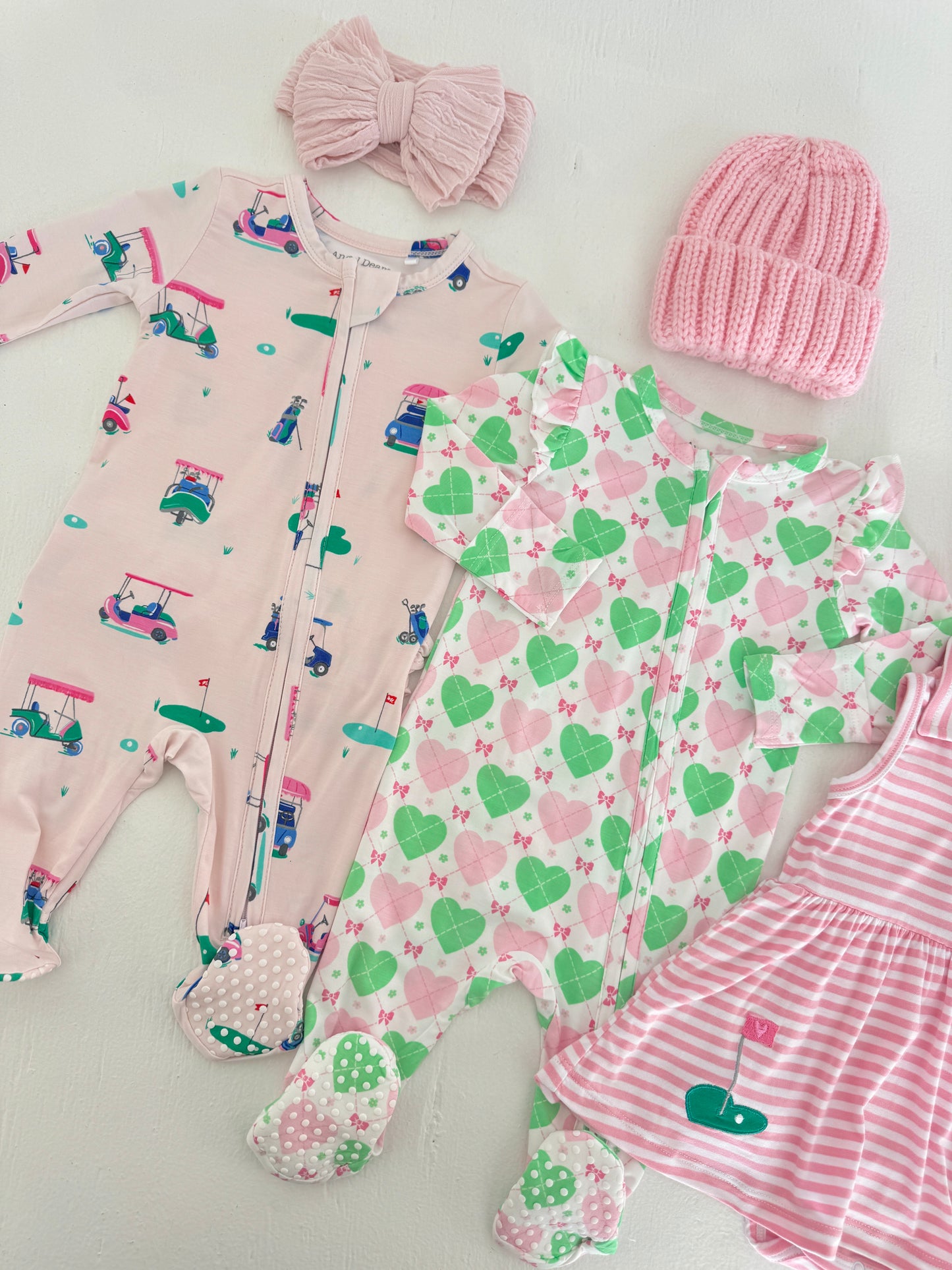 Baby outfits including two zip-up onesies, a pink headband, and a knitted hat, all with playful patterns.