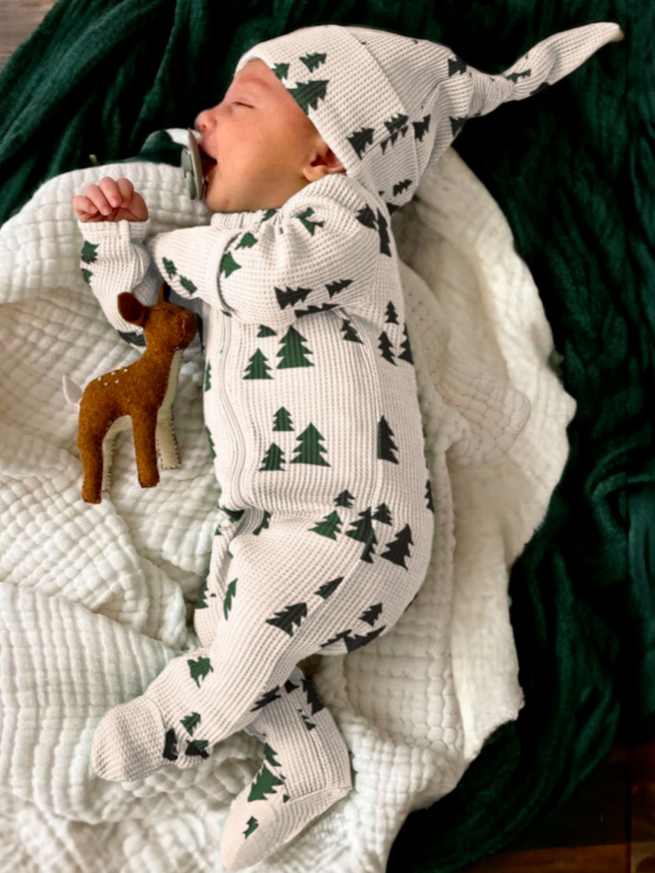 Baby wearing a tree-patterned romper and hat, yawning and cuddling a small toy deer on a cozy blanket.