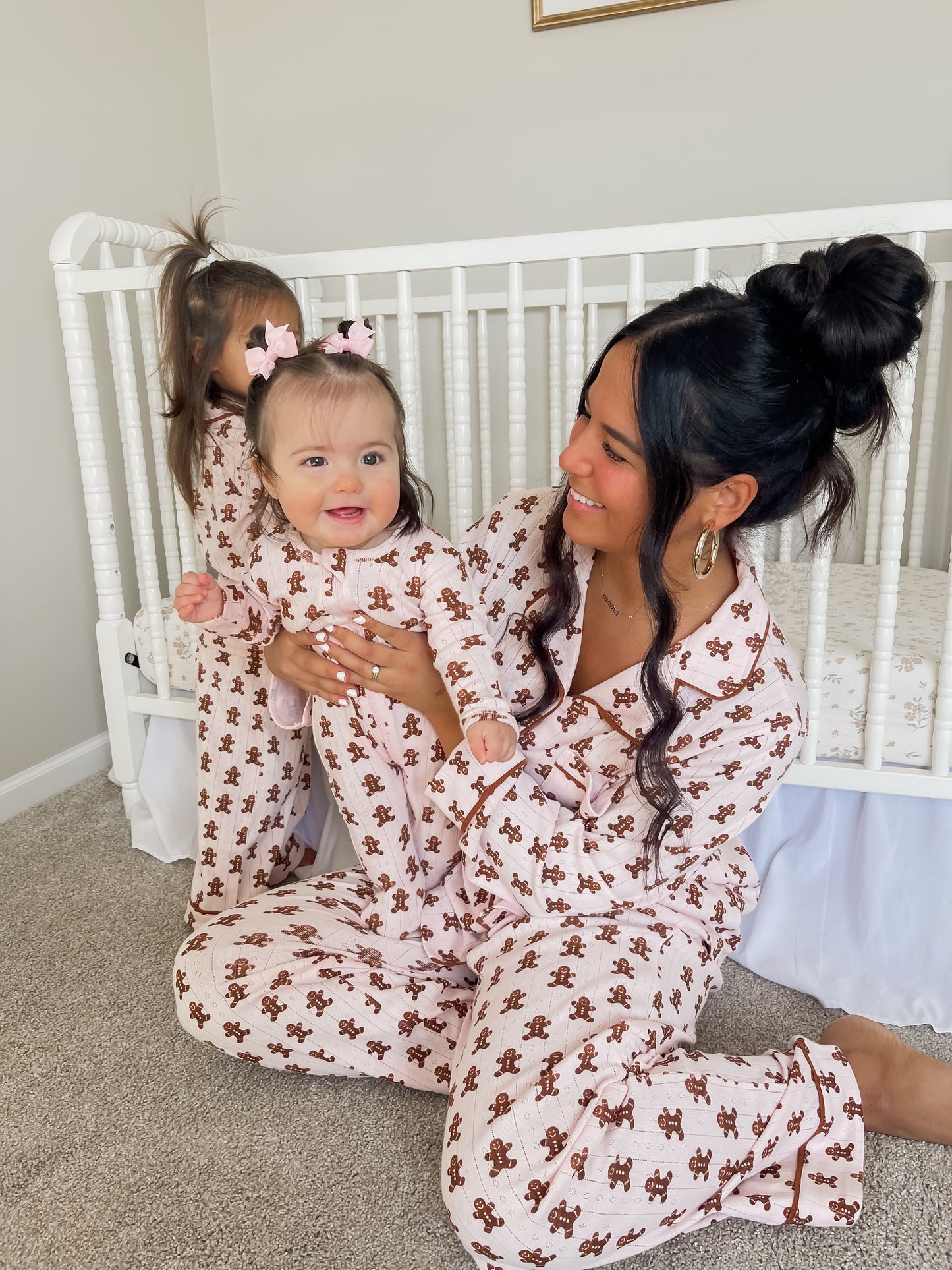 Mother and baby wearing matching pink gingerbread-themed pajamas, seated near a crib, with a playful atmosphere.