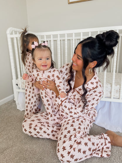 Mother and baby wearing matching pink gingerbread-themed pajamas, seated near a crib, with a playful atmosphere.