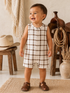 Smiling toddler in a plaid romper stands on a rug, with rustic decor in the background.