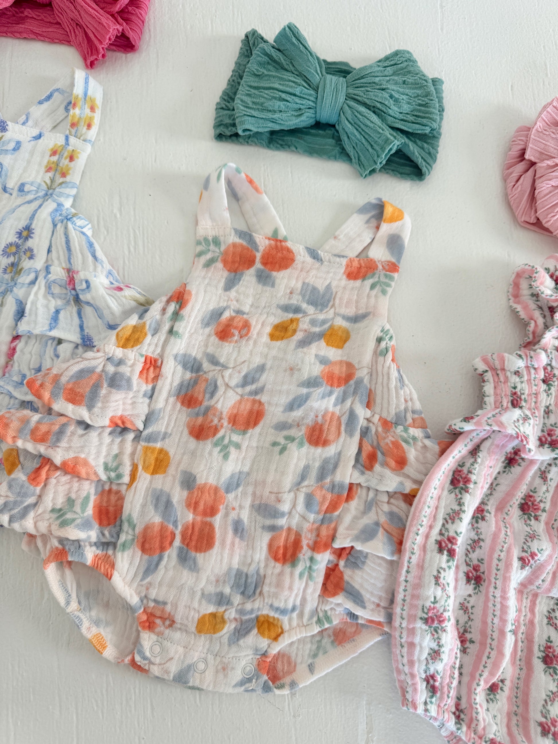 Colorful baby outfits and hair accessories on a white surface, featuring floral and fruit patterns.