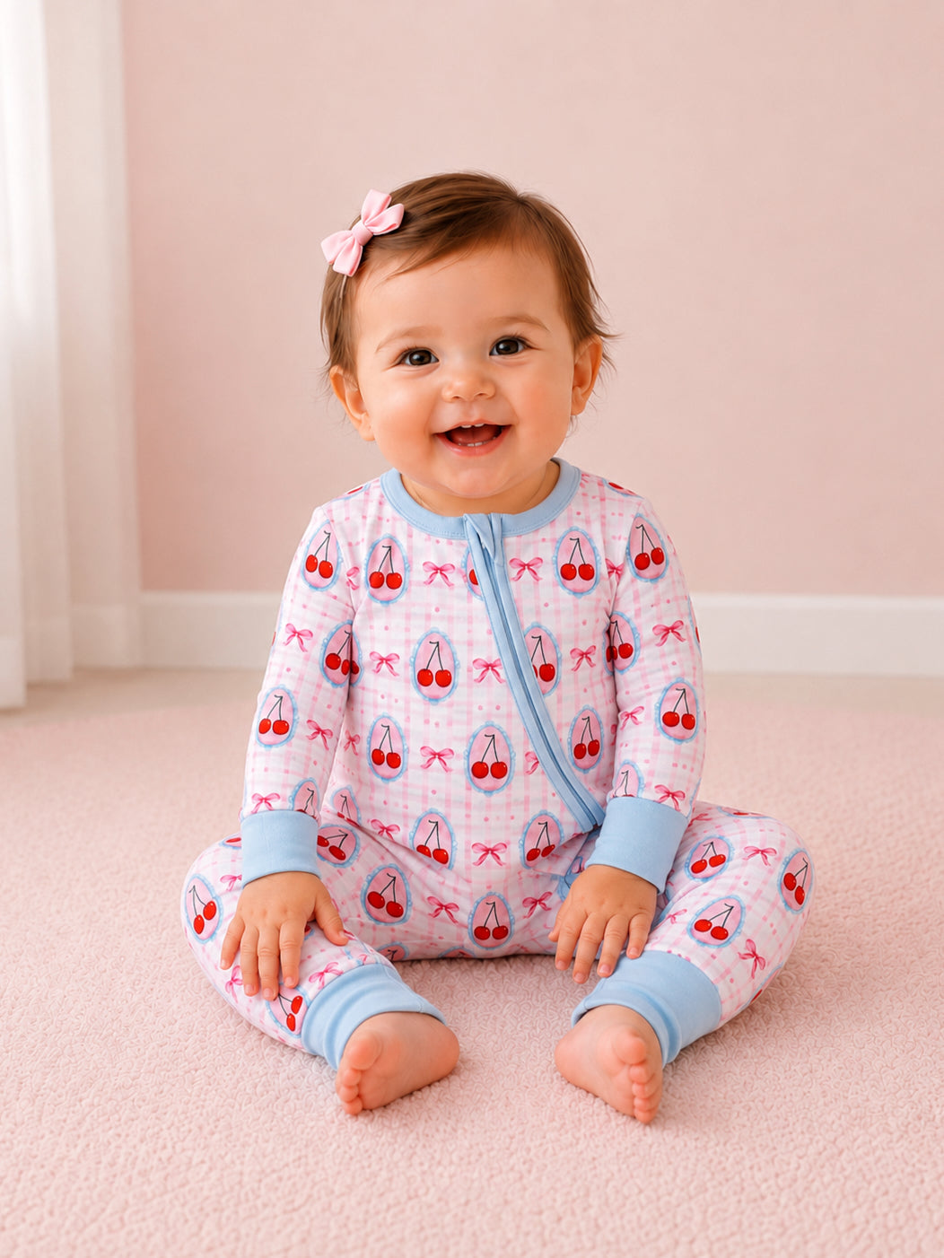 Smiling baby wearing cherry-patterned pajamas, sitting on a soft pink carpet, with a bow in their hair.