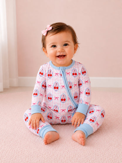 Smiling baby wearing cherry-patterned pajamas, sitting on a soft pink carpet, with a bow in their hair.