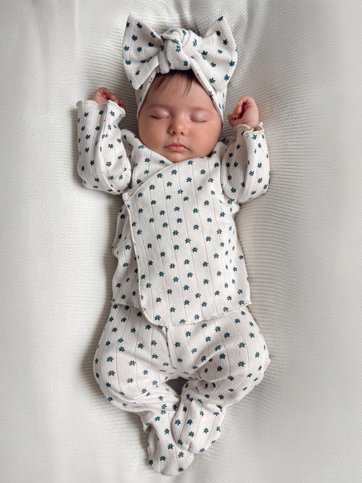 Sleeping baby in a patterned onesie with a large bow headband, resting on a textured white blanket.
