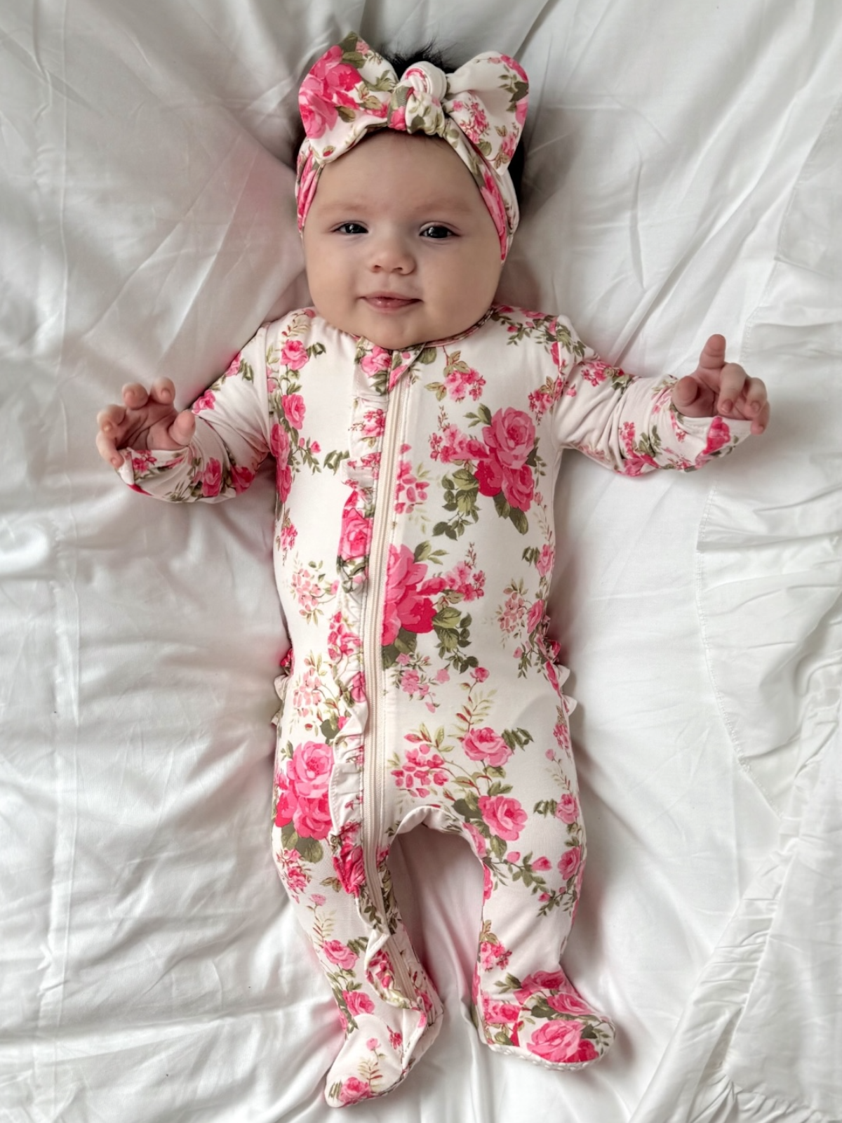 Smiling baby in floral romper and matching headband, lying on white bedding.