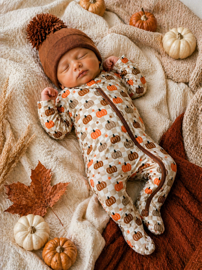 Sleeping baby in a pumpkin-patterned outfit, surrounded by autumn leaves and small pumpkins on a cozy blanket.