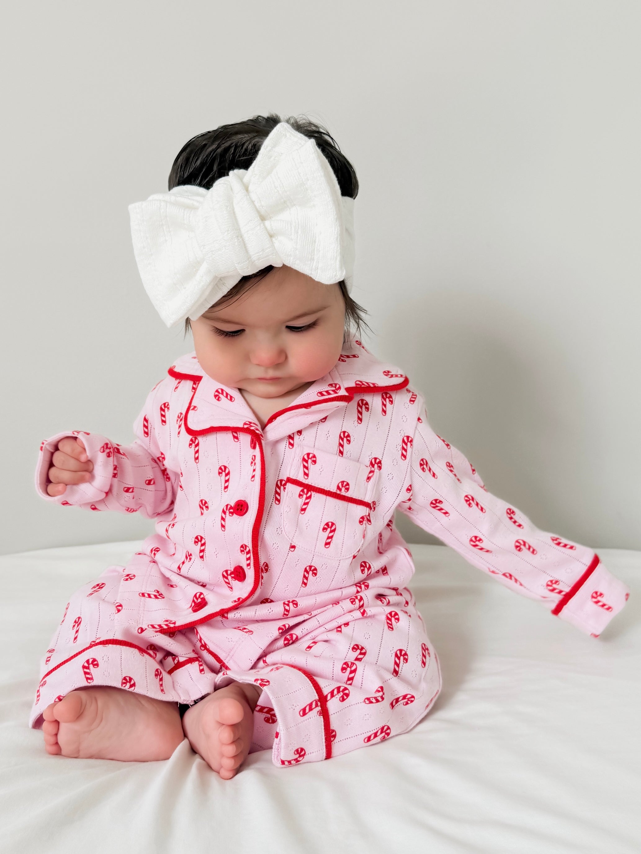Baby in pink candy cane-patterned pajamas, sitting on a bed with a large white bow headband.
