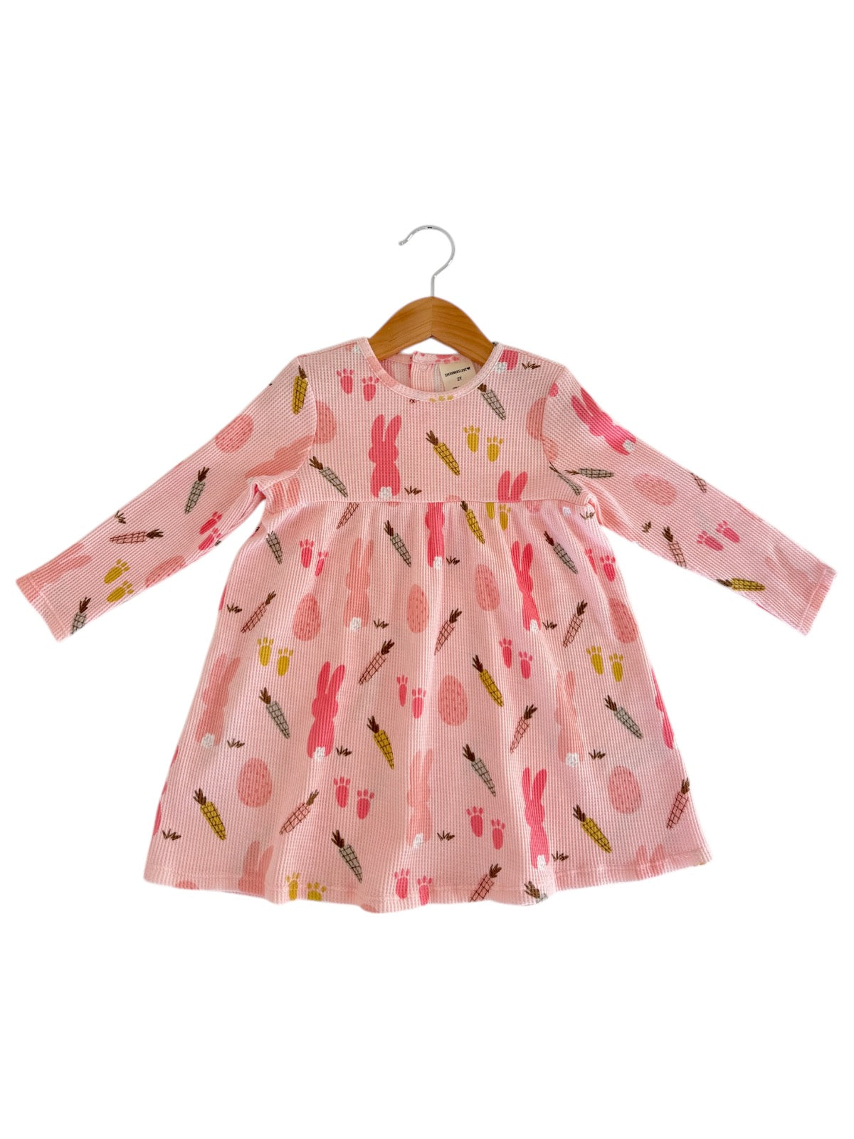 Pink long-sleeve dress with bunny, egg, and carrot prints, hanging on a wooden hanger against a white background.