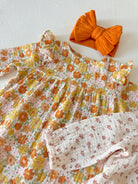 Floral baby dress with ruffled collar and a matching orange bow on a white background.