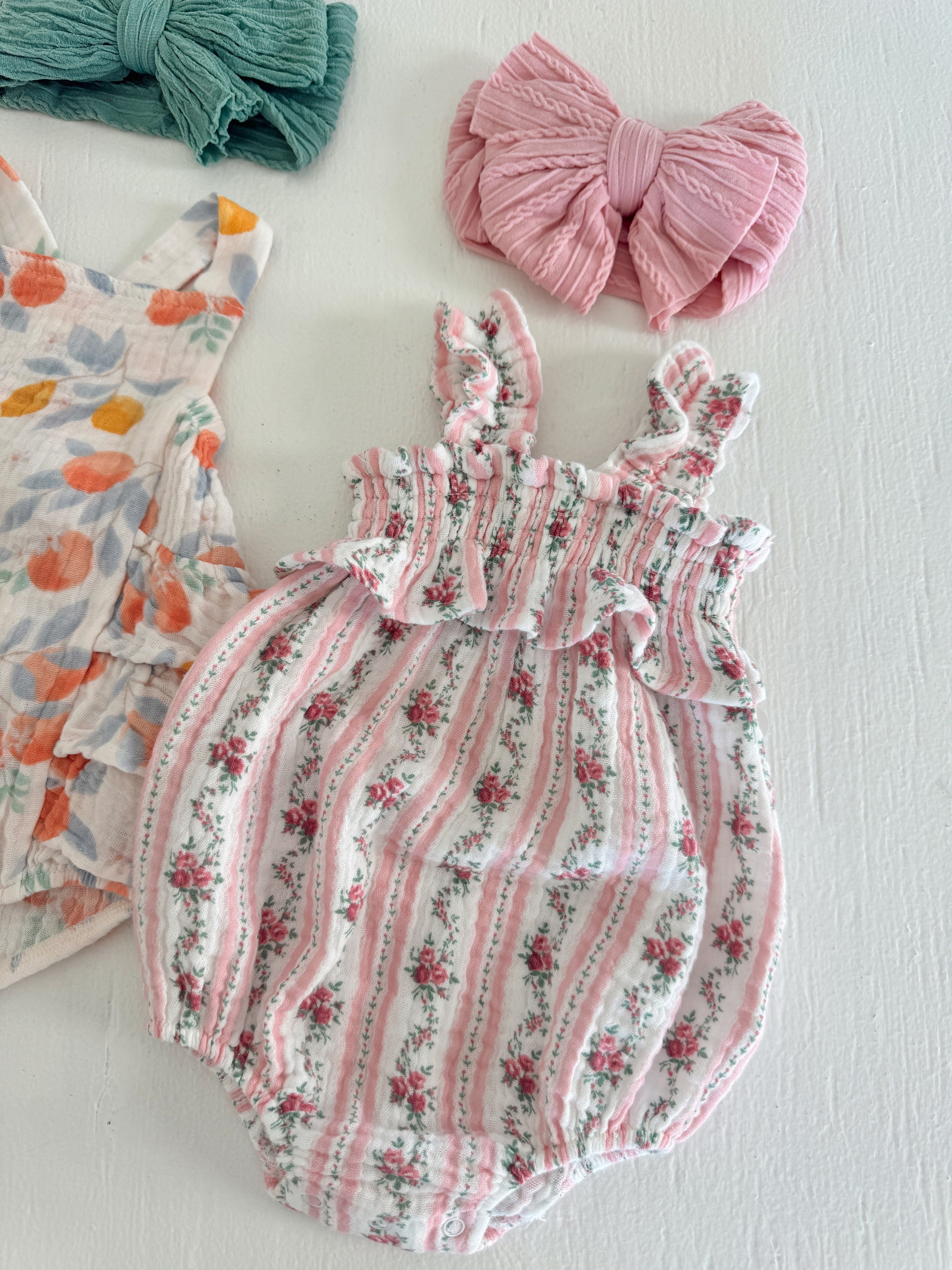 Floral baby romper with ruffled straps, paired with pink and green hair bows on a white background.