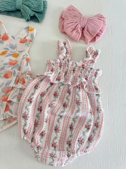 Floral baby romper with ruffled straps, paired with pink and green hair bows on a white background.