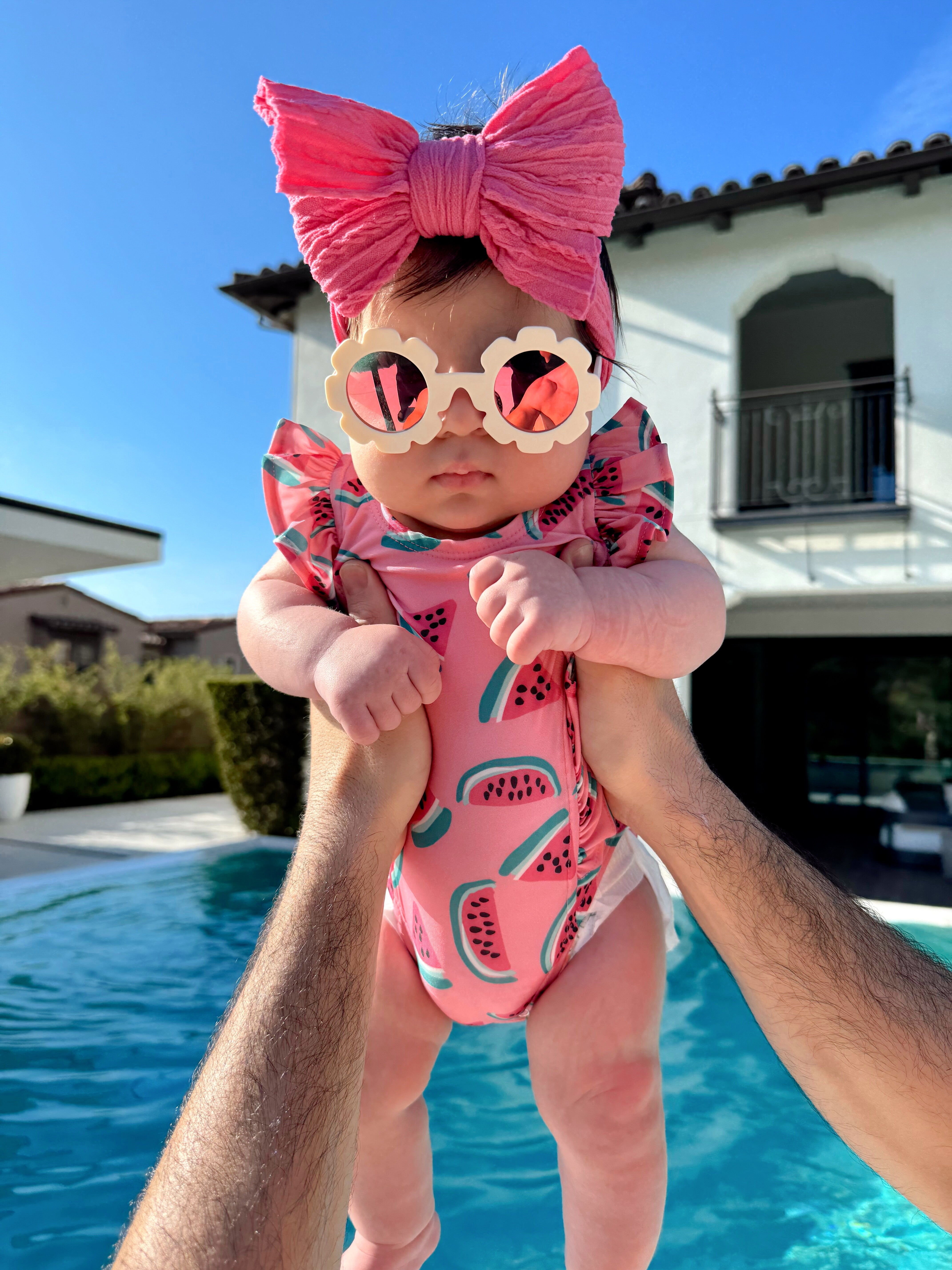Baby in a pink watermelon swimsuit and sunglasses is held above a pool, wearing a large pink bow on a sunny day.