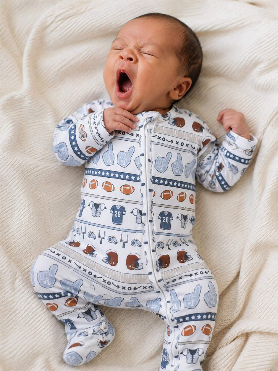 Baby in a football-themed onesie yawning on a cozy blanket backdrop.