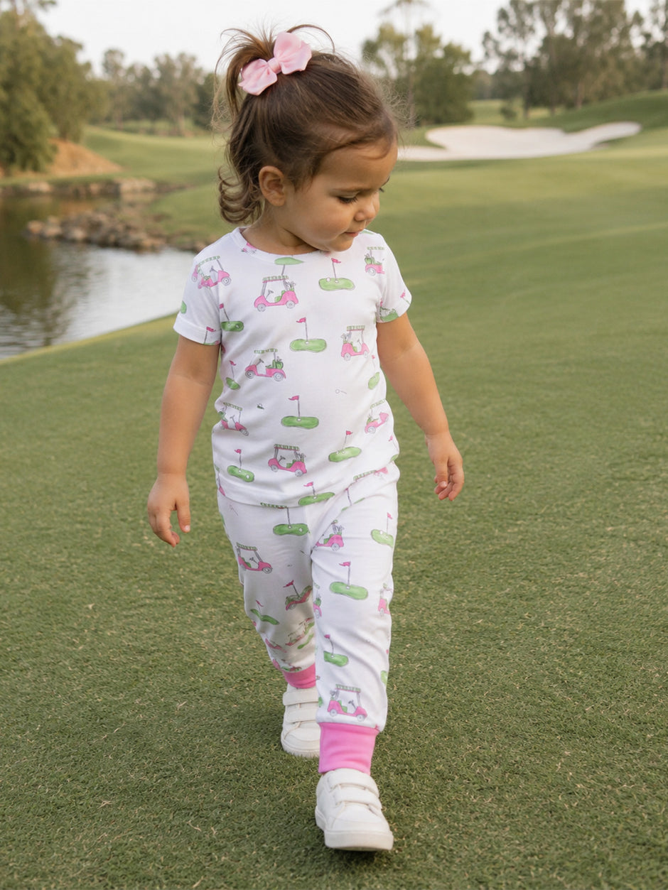 Child in golf-themed pajamas walks on grass near a lake, with trees and a golf course in the background.