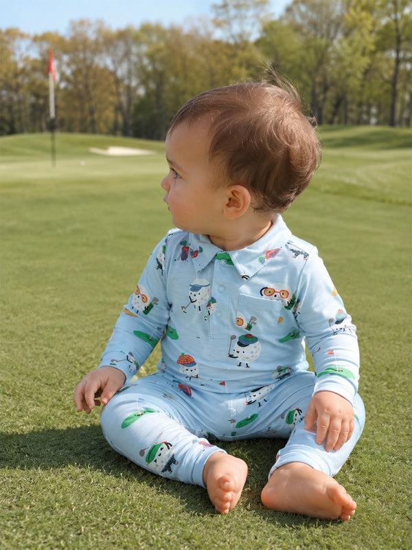 Child in playful outfit sits on green grass outdoors, looking to the side with a golf course backdrop.