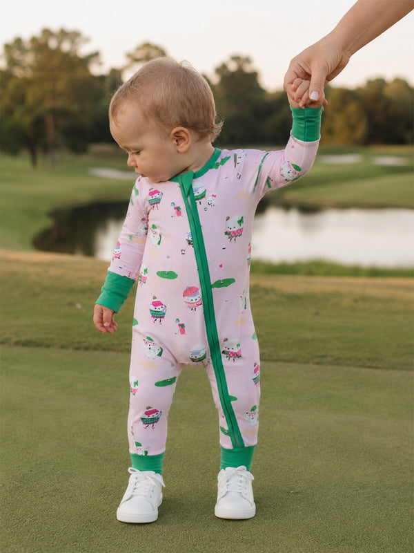 Toddler in colorful ice cream-themed onesie, holding an adult's hand on a green outdoor surface.