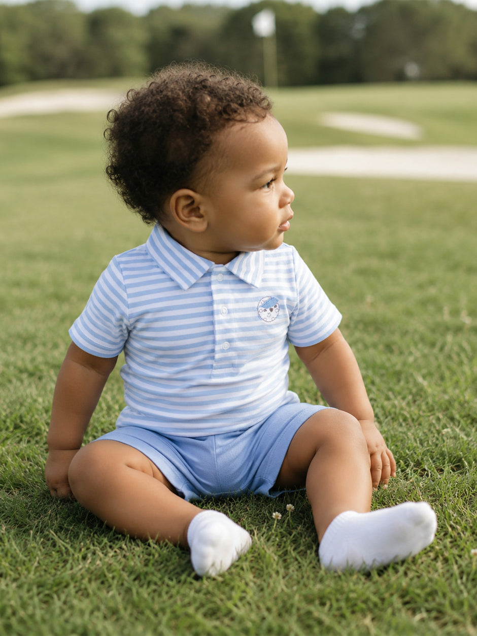 Child in blue striped polo and shorts sitting on green grass at a golf course, looking thoughtfully to the side.