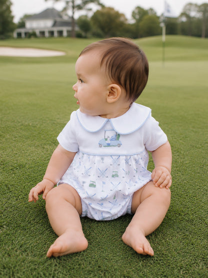 Baby sitting on green grass, wearing a white outfit with a golf-themed design, looking to the side.