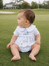 Baby sitting on green grass, wearing a white outfit with a golf-themed design, looking to the side.