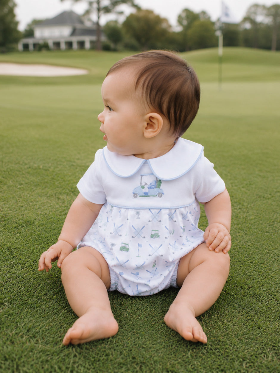 Baby sitting on green grass, wearing a white outfit with a golf-themed design, looking to the side.