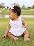 Toddler sitting on grass in a floral dress with a golf cart design, wearing a pink bow headband.