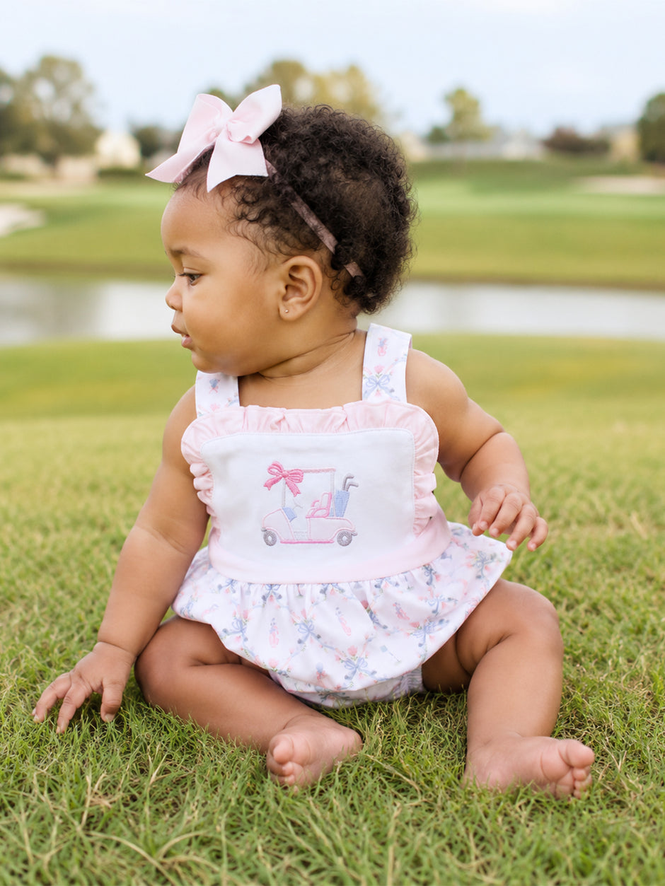 Toddler sitting on grass in a floral dress with a golf cart design, wearing a pink bow headband.