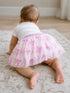 Baby girl crawling on a rug, wearing a white shirt and a pink bow-patterned tutu.