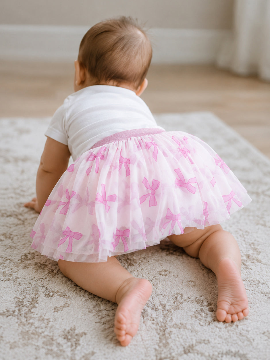 Baby girl crawling on a rug, wearing a white shirt and a pink bow-patterned tutu.