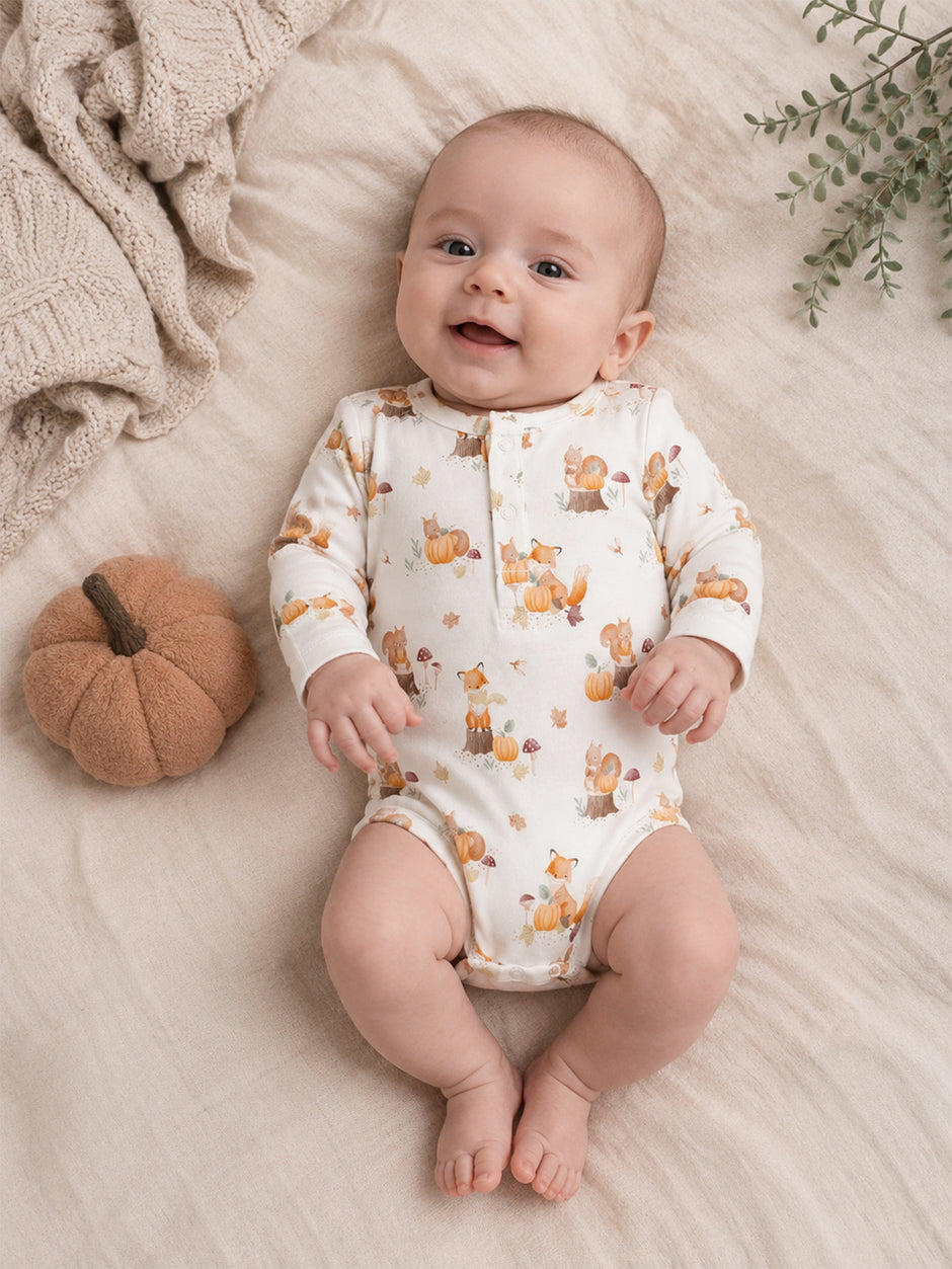 Smiling baby in a printed onesie with foxes, pumpkins, and leaves, resting on a soft blanket. Cozy autumn vibe.