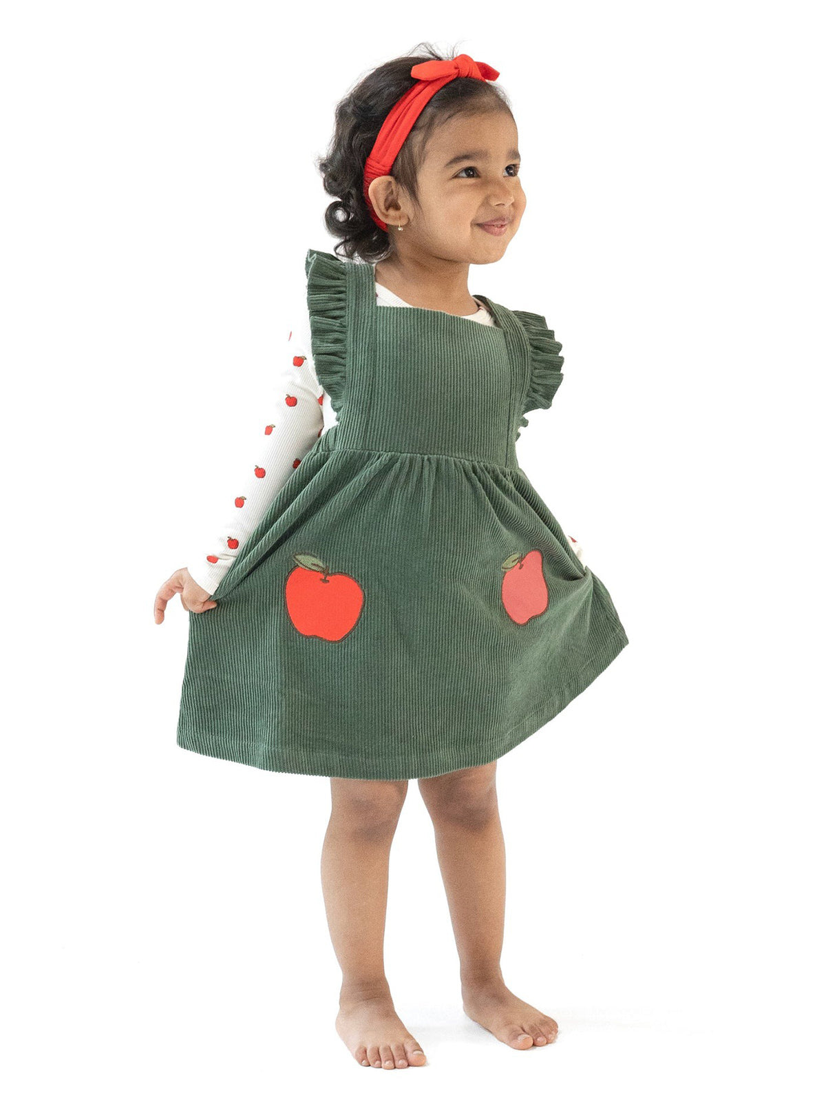Smiling toddler in a green corduroy dress with apple patches, wearing a red headband and long-sleeve top with hearts.