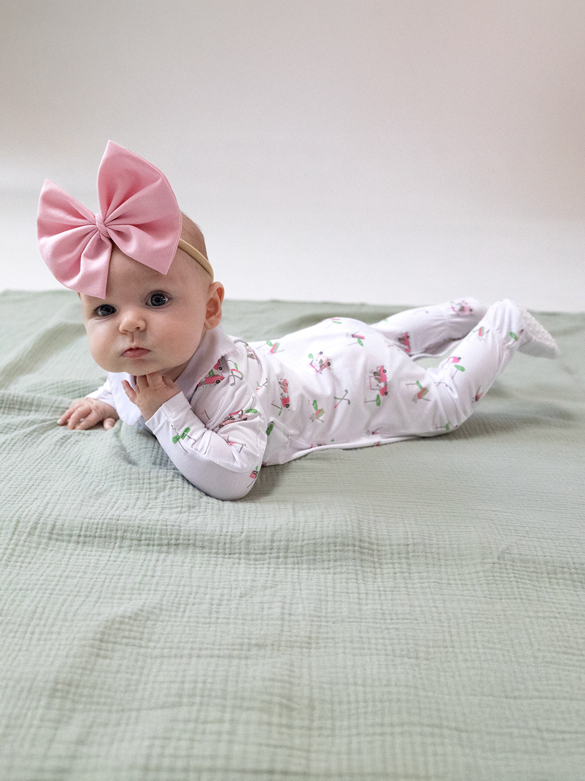 Baby laying on a green blanket, wearing a white outfit with colorful patterns and a large pink bow on her head.