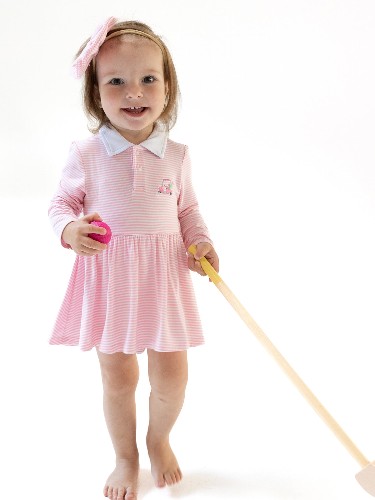 Smiling child in a pink striped dress holding a toy and a wooden stick, standing on a plain white background.