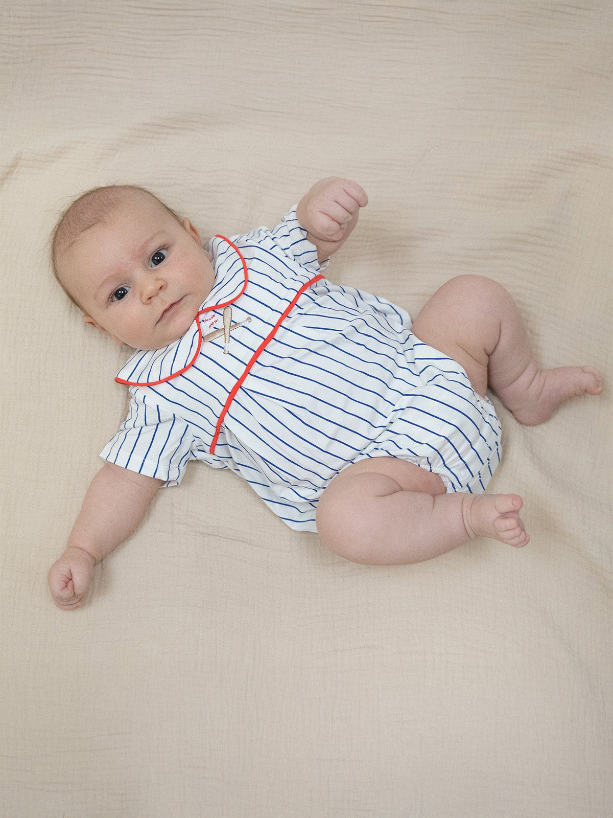 Baby in a striped romper with red trim, lying on a textured beige surface, looking directly at the camera.