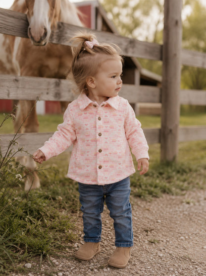 Toddler in a pink shirt and jeans stands by a wooden fence, with a horse in the background.