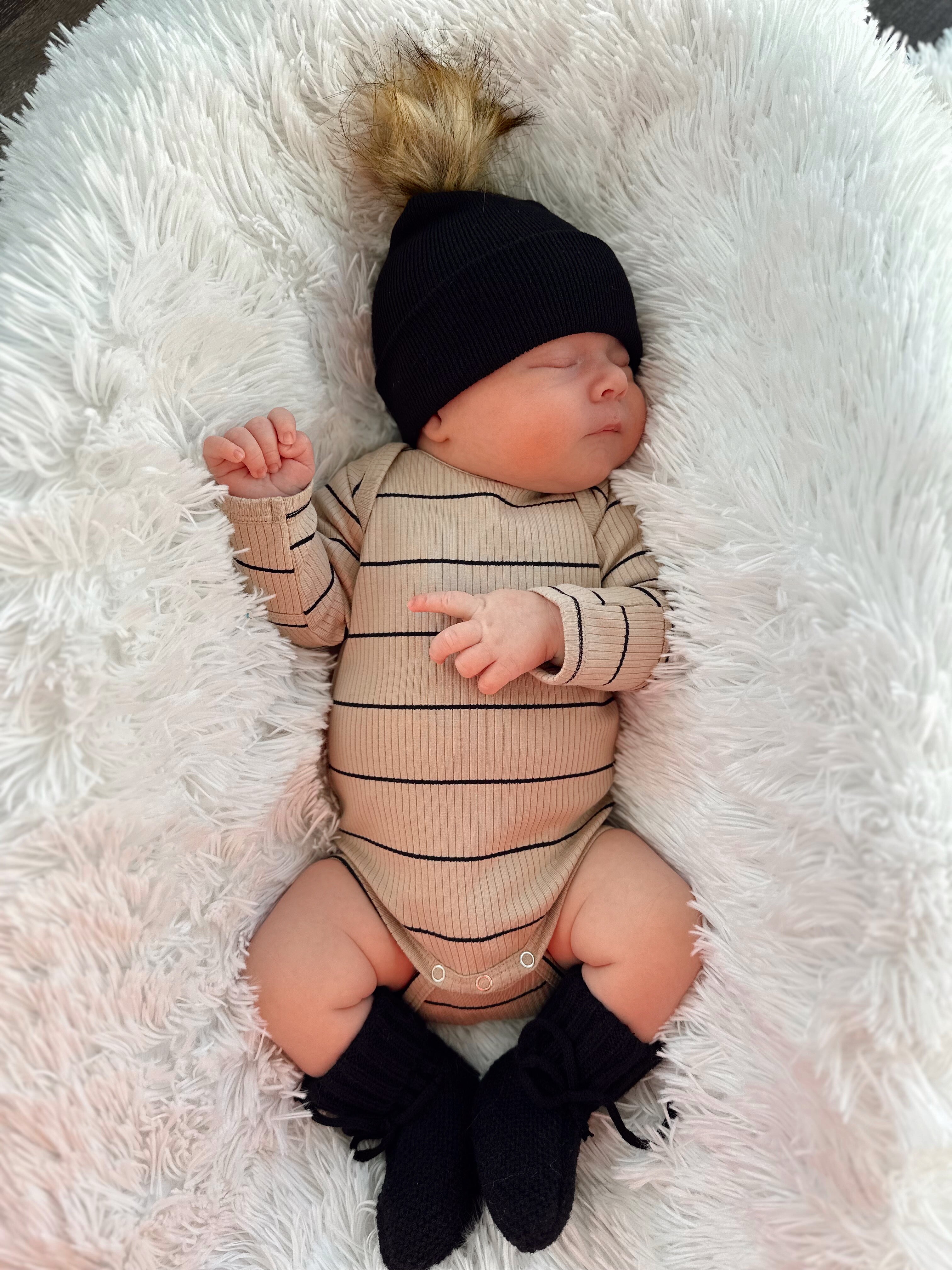 A sleeping baby in a striped onesie and black beanie, resting on a fluffy white blanket.