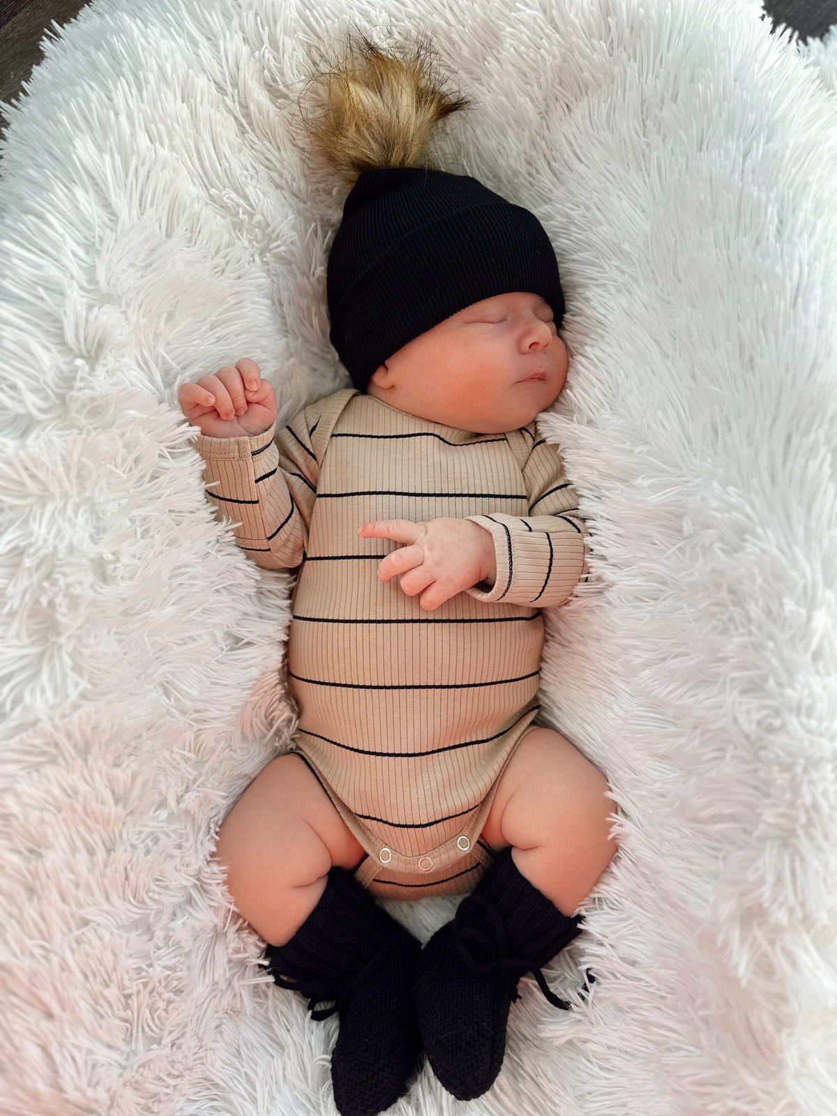 A sleeping baby in a striped onesie and black beanie, resting on a fluffy white blanket.