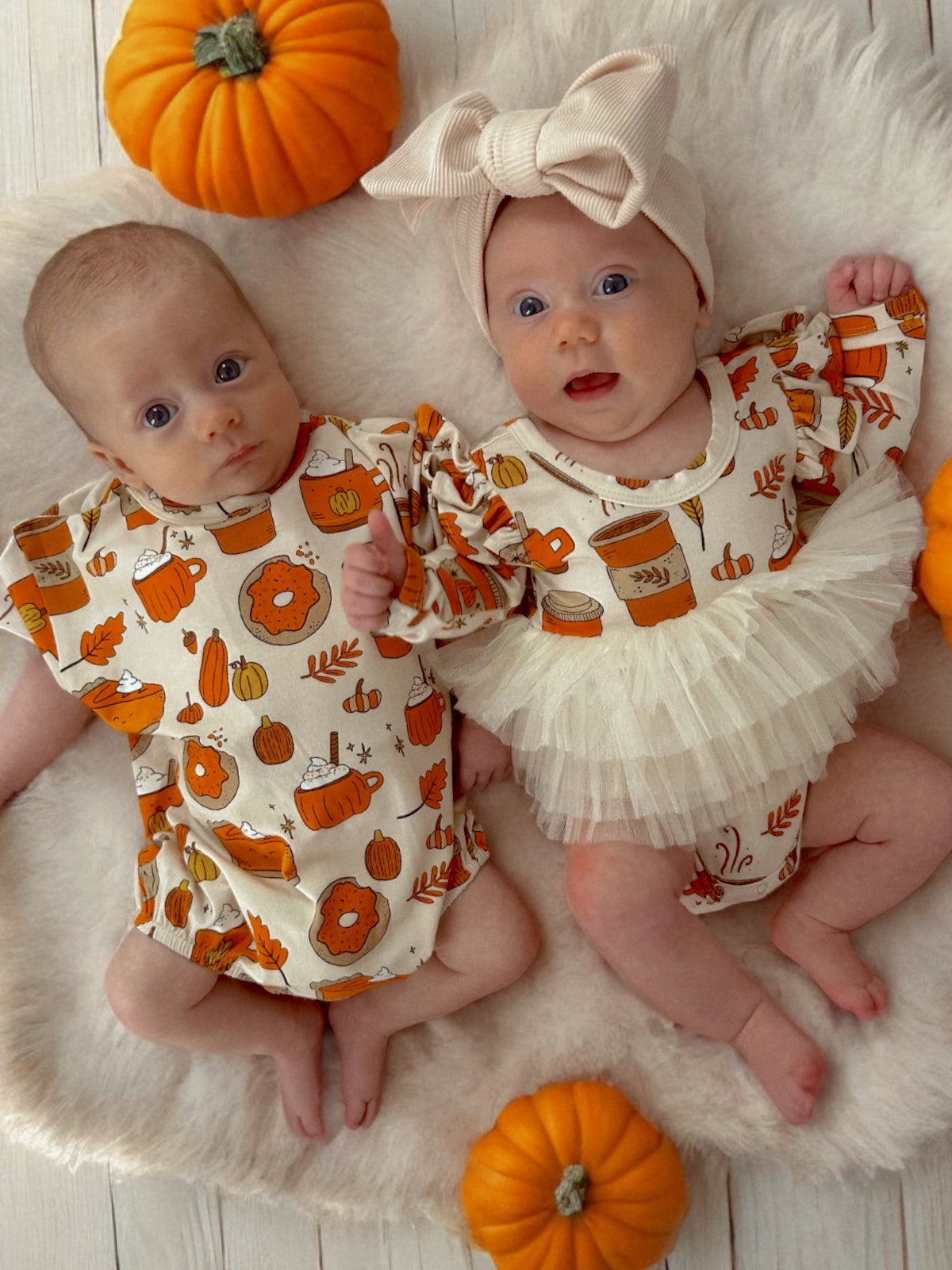 Two babies in autumn-themed outfits sit on a fluffy rug surrounded by pumpkins, smiling and posing for the picture.