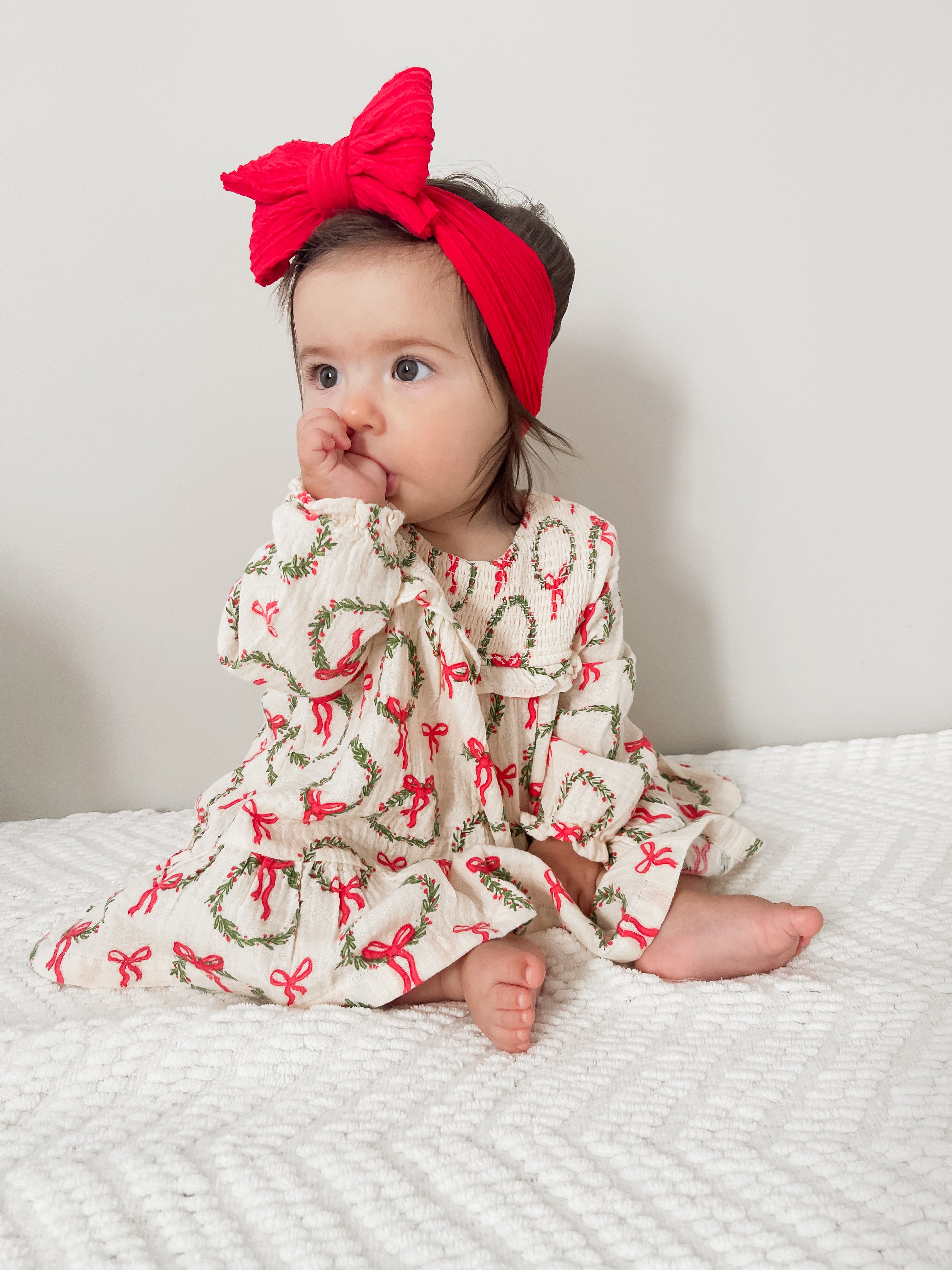 Baby girl sitting on a textured blanket, wearing a festive dress with wreaths and a bright red bow headband.