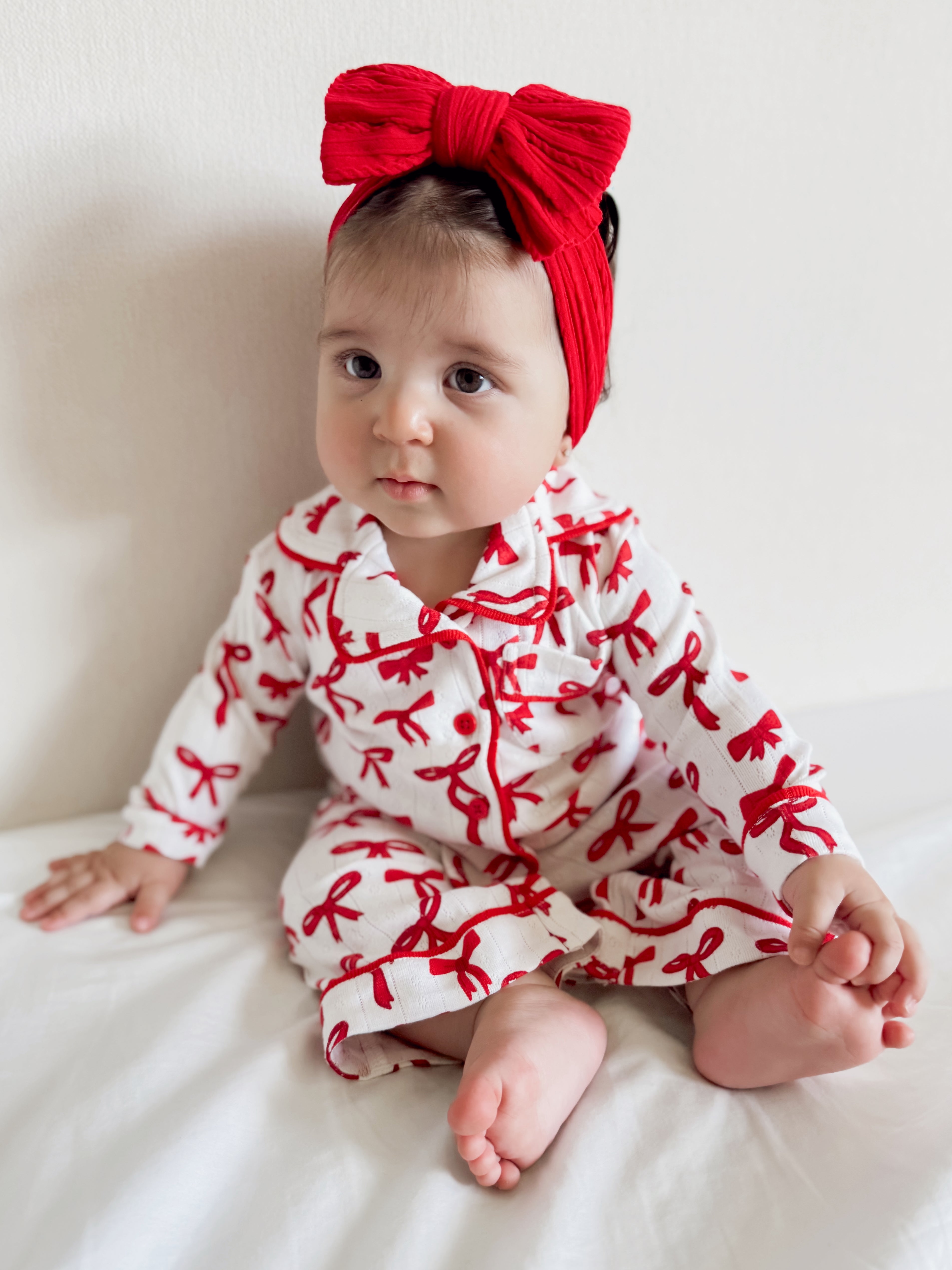 Baby girl sitting on a bed, wearing a white outfit with red bow patterns and a large red headband.