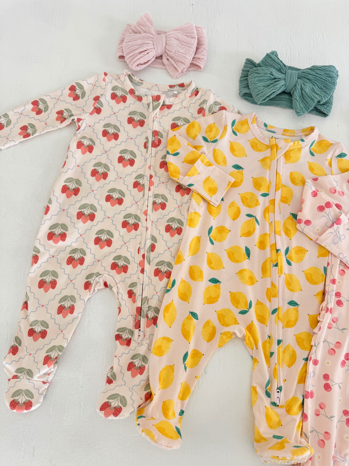 Two infant onesies with strawberry and lemon patterns, accompanied by pink and green fabric headbands.