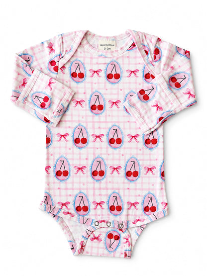 Pink baby onesie with cherry print and bows, featuring long sleeves and snap closures at the bottom.